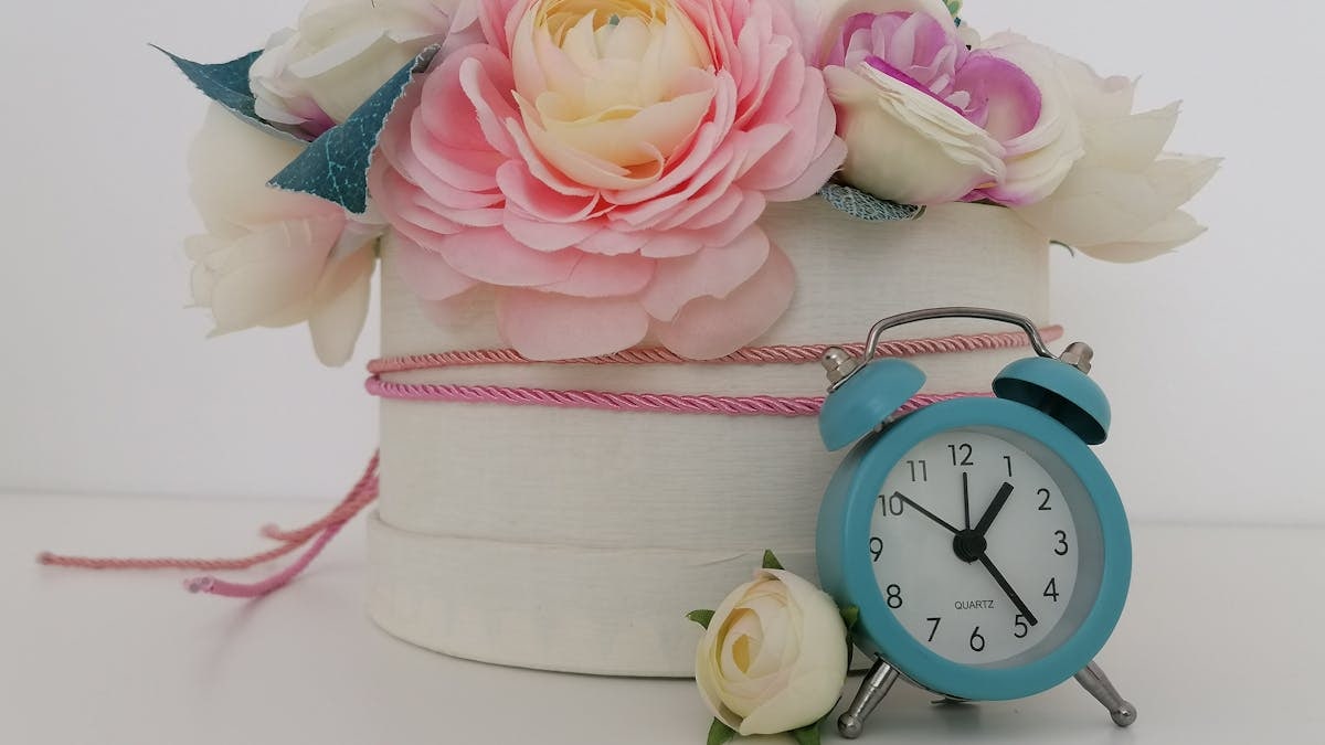 Pastel artificial flowers in vase with a vintage-style blue clock on a white background.
